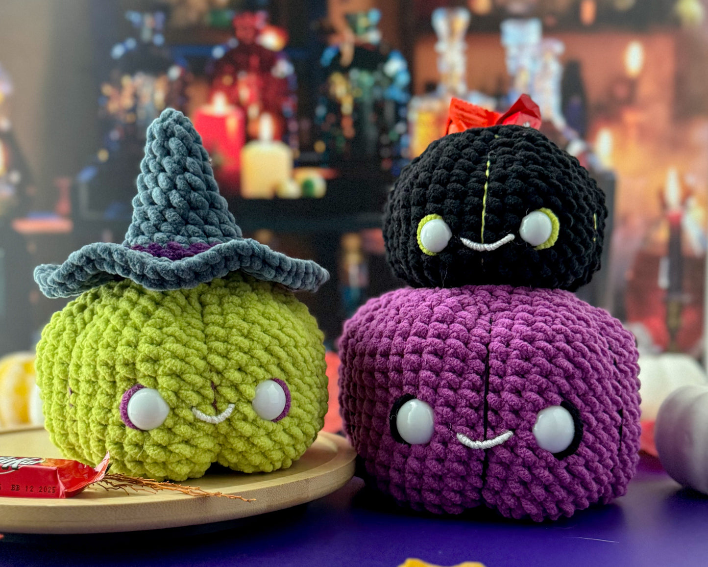 Three crocheted Halloween-themed pumpkins on a table with a blurred background of bottles and candles.