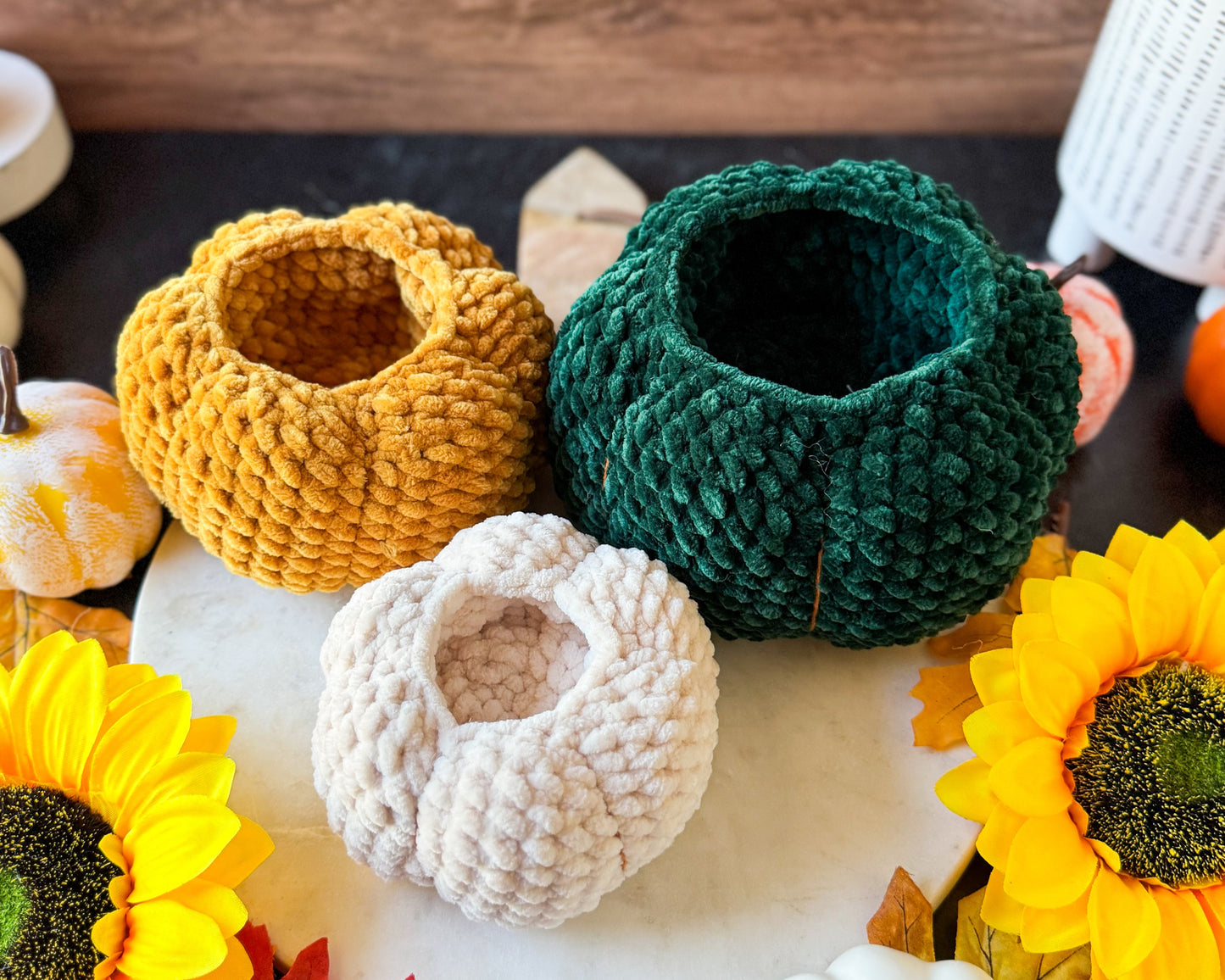 Three crochet pumpkin bowls in yellow, green, and white on a wooden surface with decorative elements.