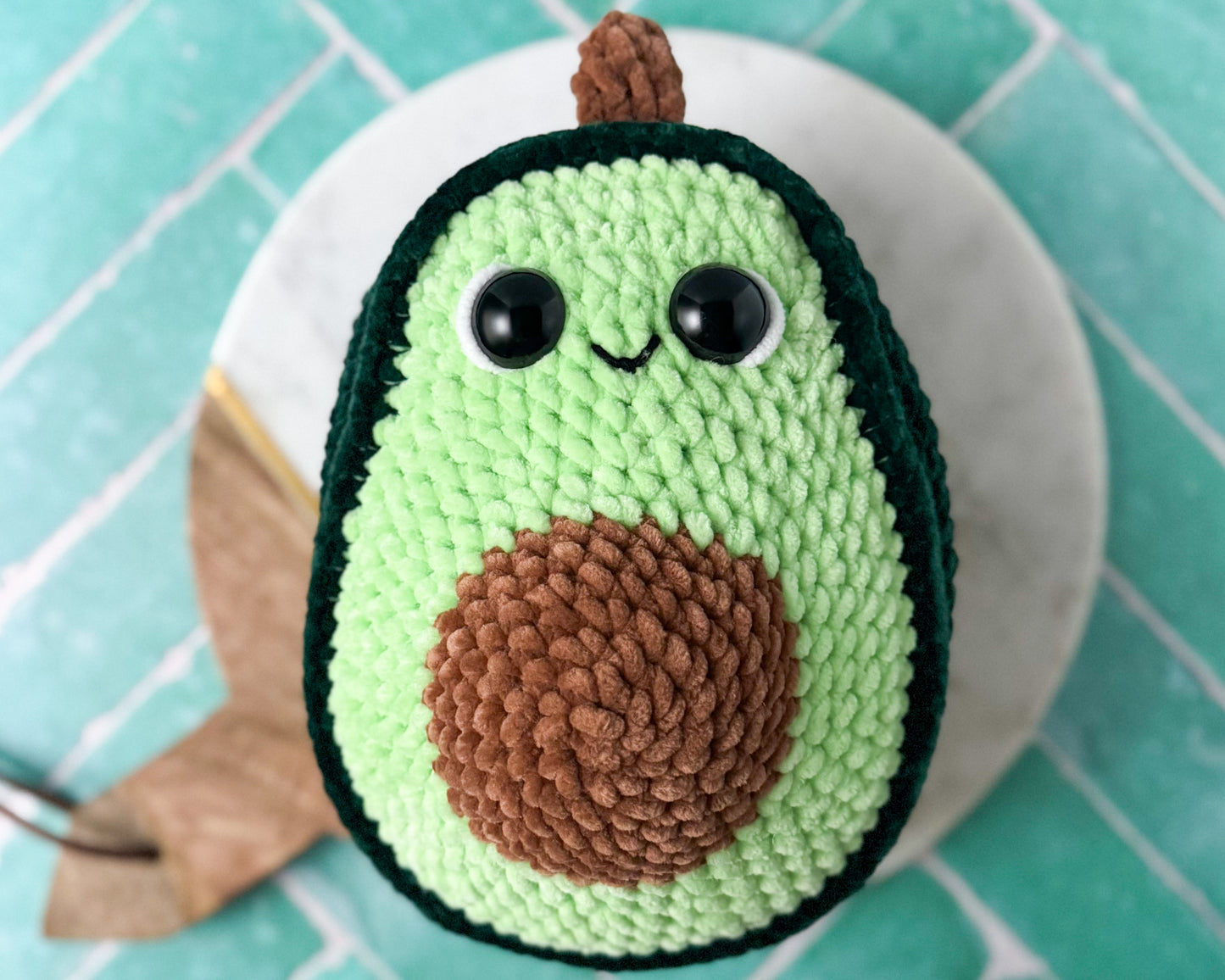 Crocheted avocado plush toy on a white plate with a turquoise tiled background