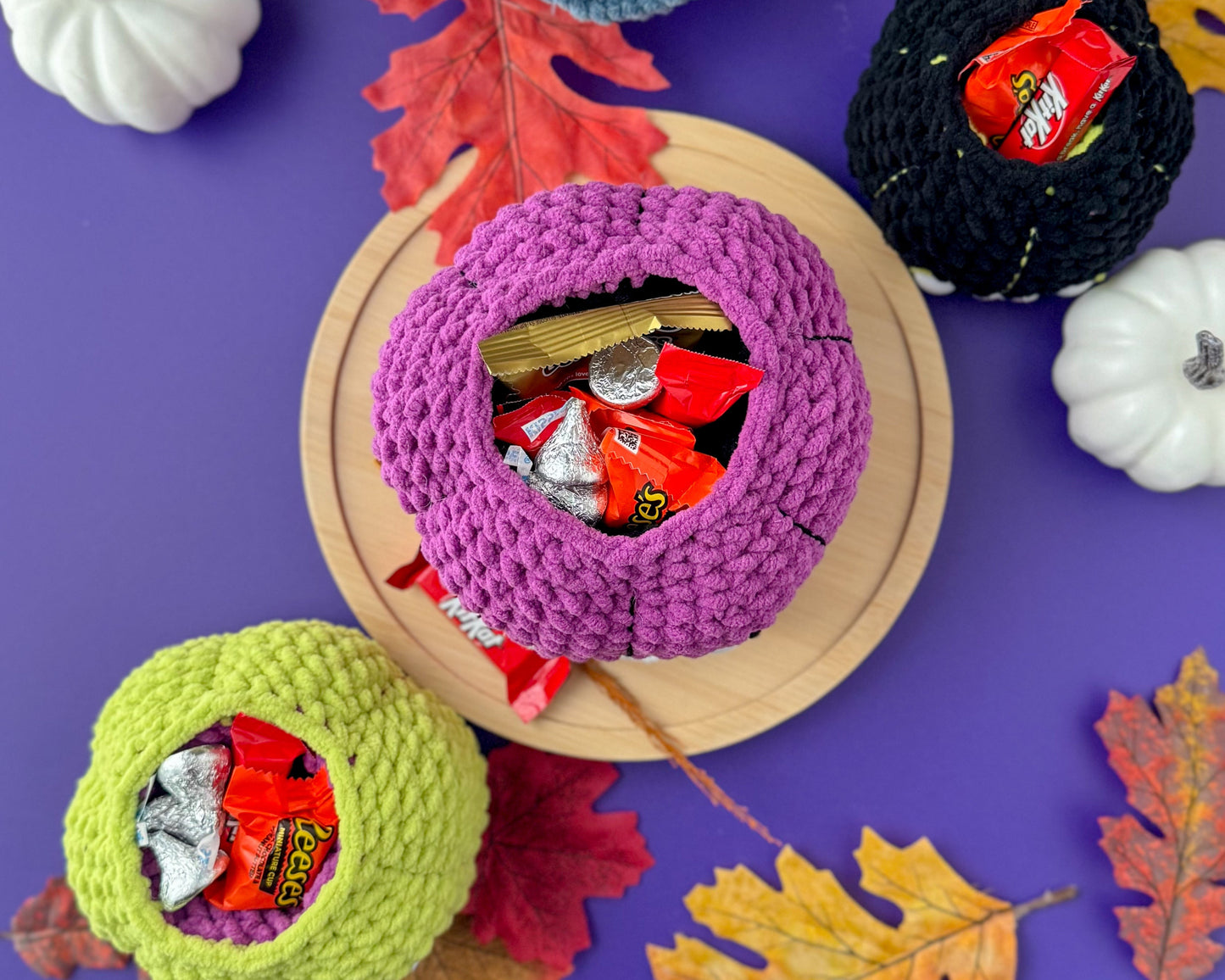 Colorful crochet pumpkin bowls filled with candy on a purple background with pumpkins and leaves.