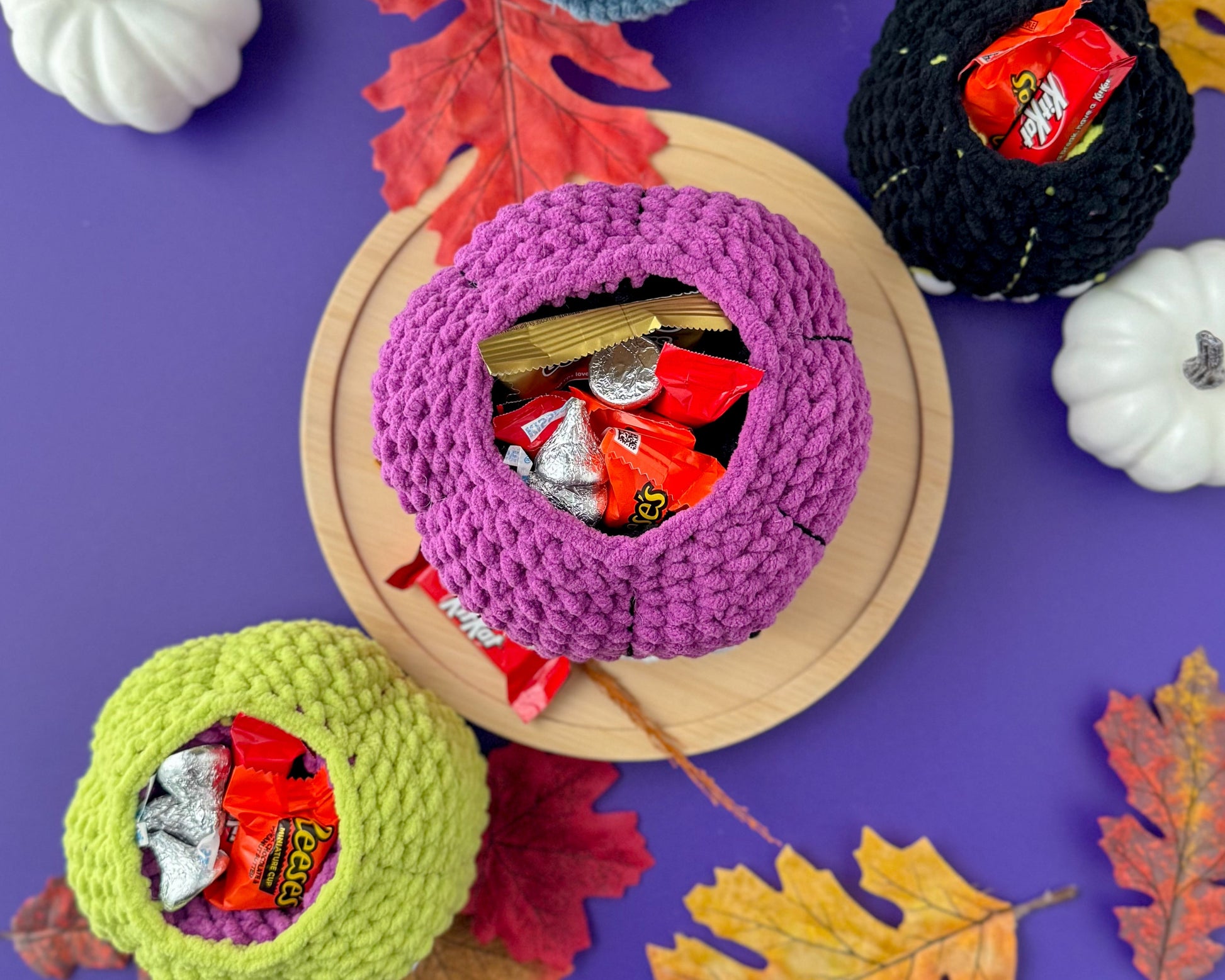 Colorful crochet pumpkin bowls filled with candy on a purple background with pumpkins and leaves.