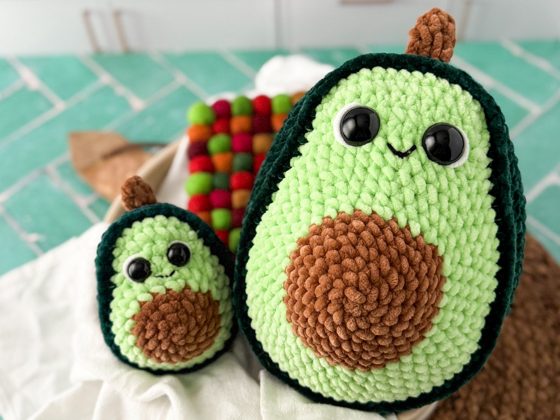 Two crocheted avocado toys on a green surface with colorful items in the background.