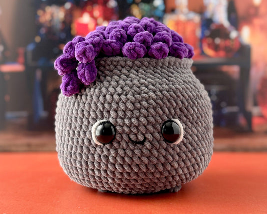 A cute, round gray crocheted plush cauldron toy with a purple potion and large glossy eyes, sitting on an orange surface with a blurred background.