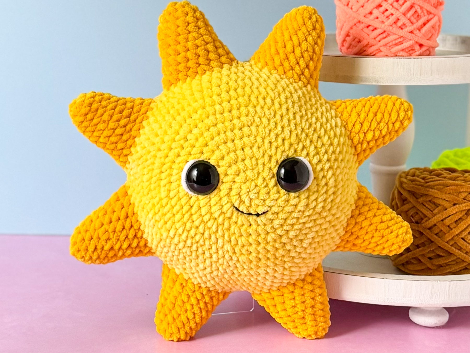 Crocheted sun-shaped toy with black eyes and a smile on a pink surface with yarn balls in the background.