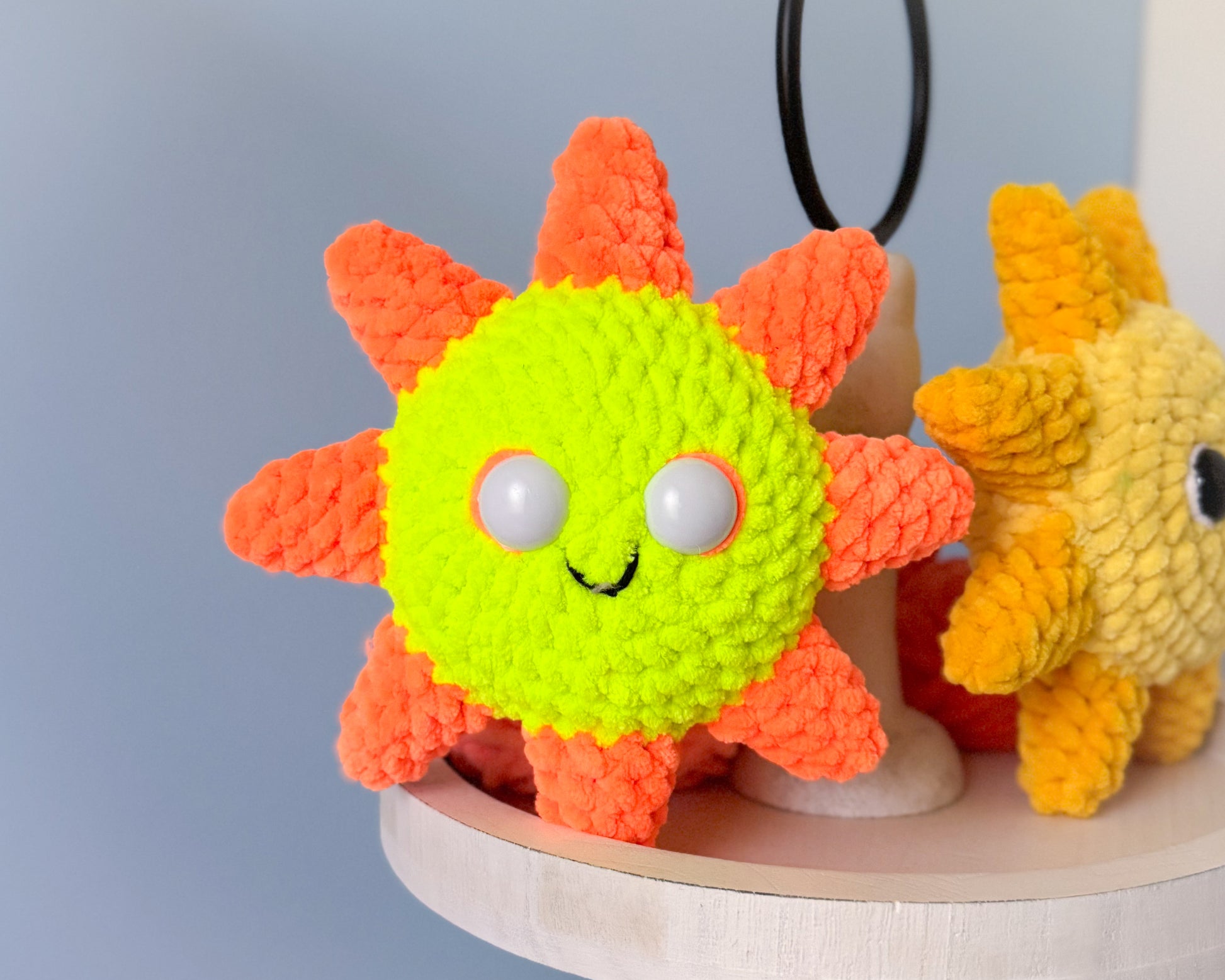 Two knitted or crocheted sunshines, one neon yellow  and orange, the other yellow, on a blue gray background.