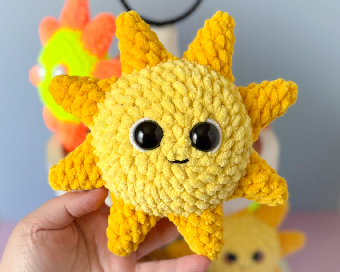 Hand holding a yellow crochet sun-shaped toy with black eyes and a small mouth against a blurred background.