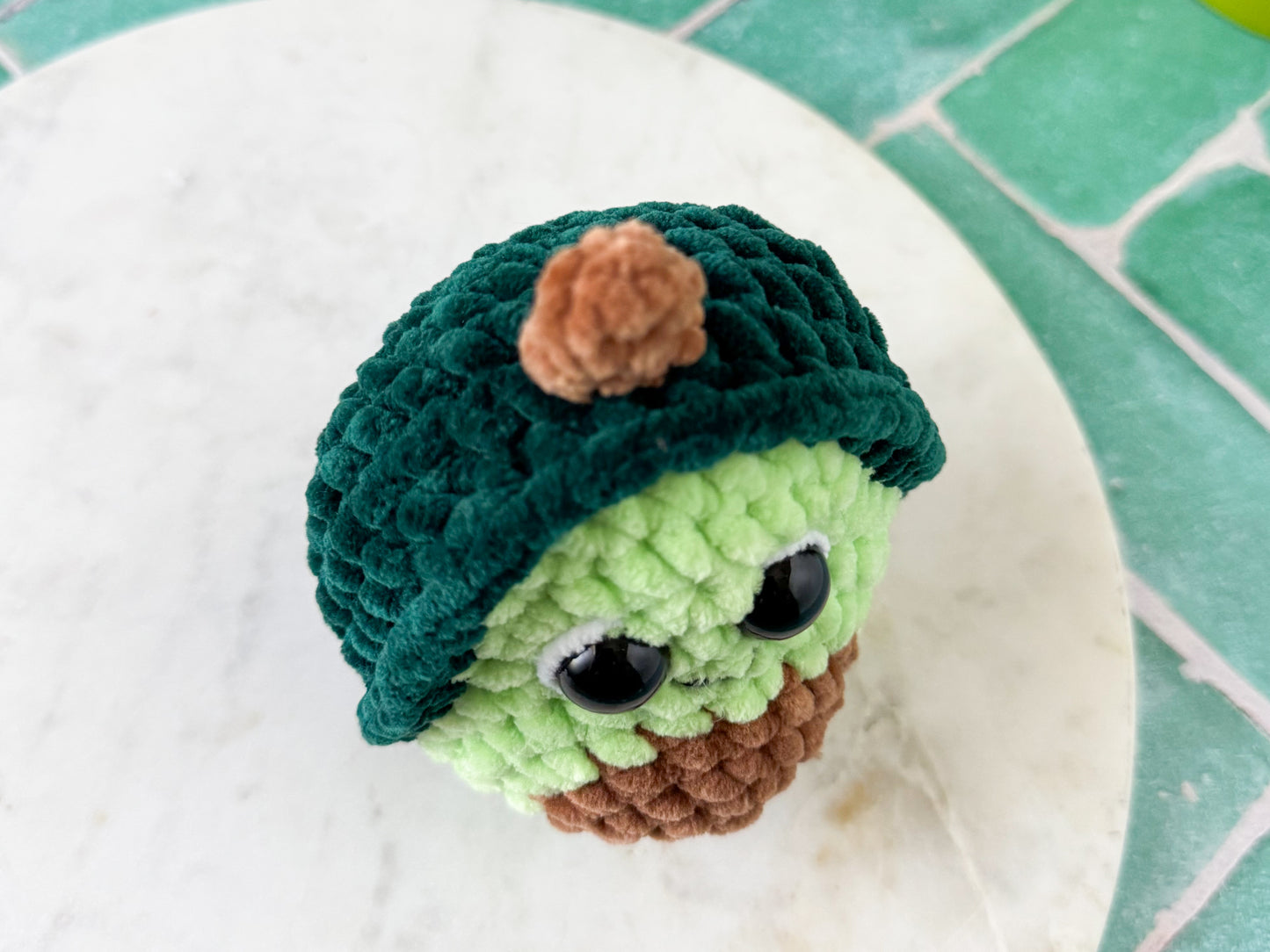 Small crochet avocado on a white surface with a green tiled background