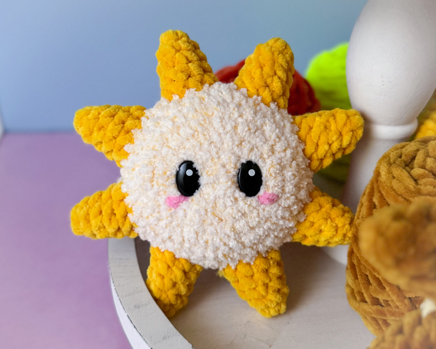 Crocheted sun-shaped plush toy with black eyes and pink cheeks on a light surface.