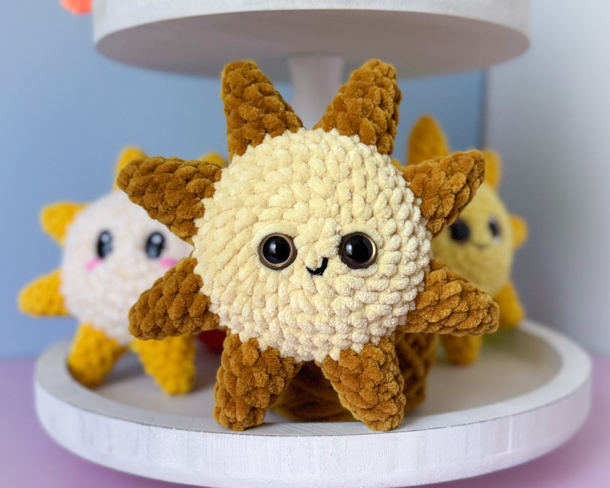 Three small plush toys resembling a sunshine on a white stand with a light blue background